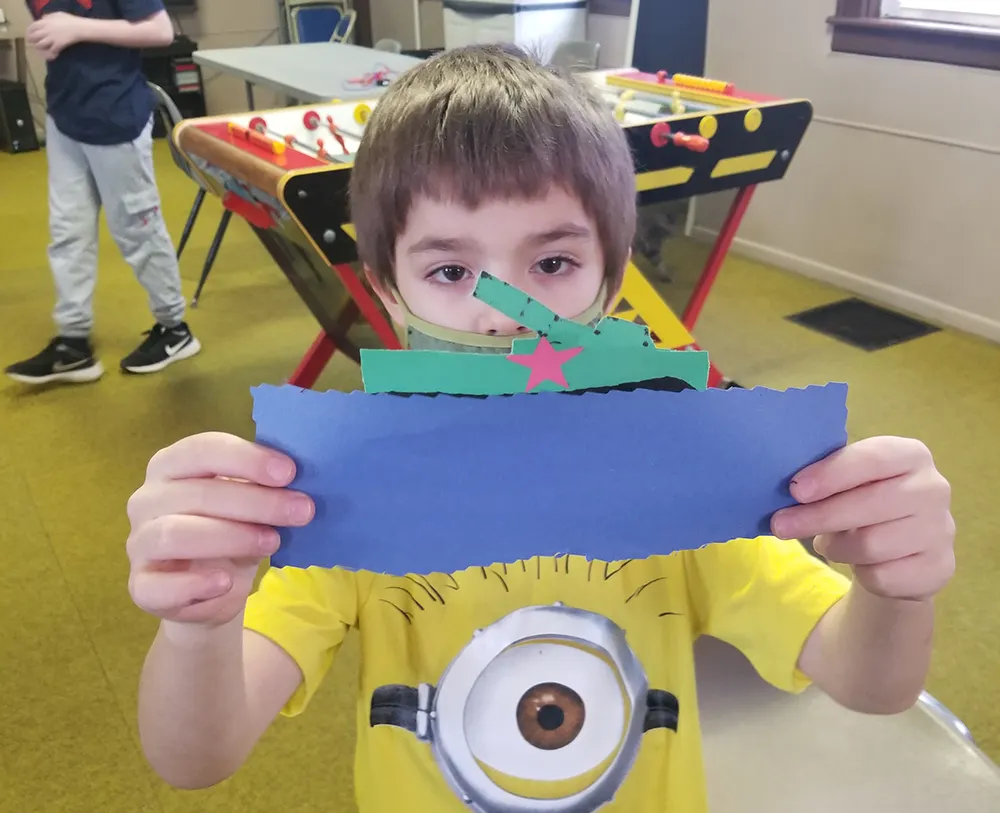 A YA kid holds up a paper art tank he made from construction paper