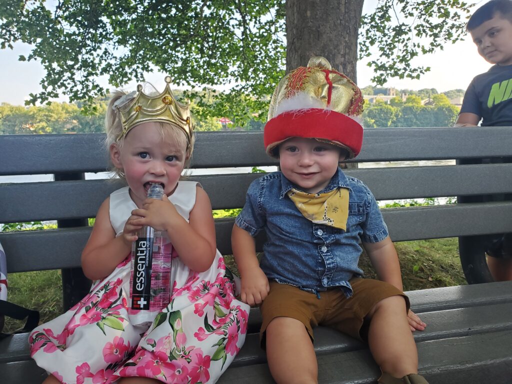 Two toddlers dressed as a king and a queen on a beach smile outside on a sunny day
