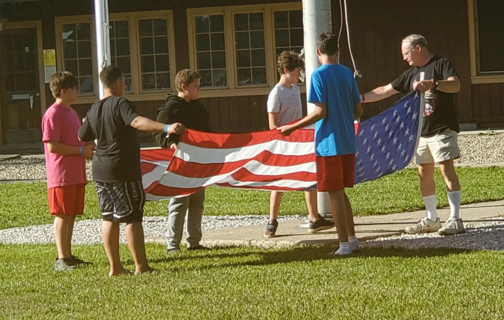 5 Children help a camp counselor adult fold the American flag next to its pole after it was taken down for the day.