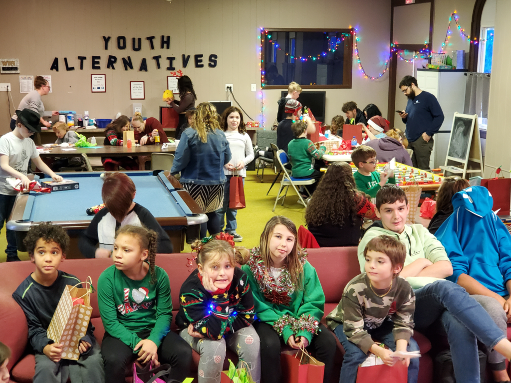 Children in Christmas clothing sitting on a couch inside YA while kids stand and enjoy a Christmas party in the background