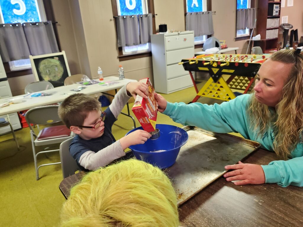 A YA Staff member helps a child pour baking ingredients in a bowl at YA