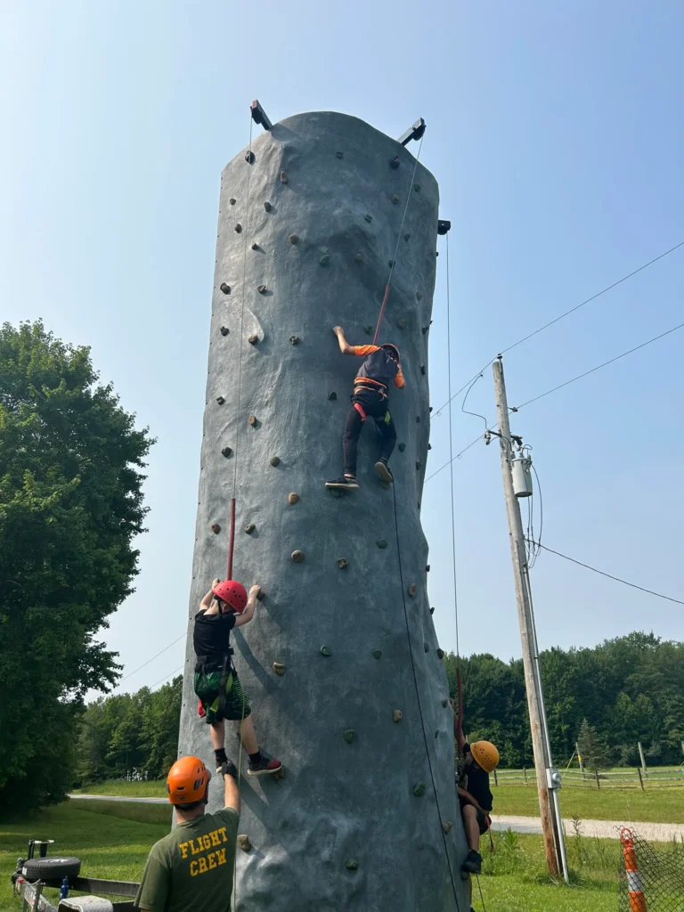 YA kids climb a cylindrical artificial rock climbing tower, wearing harnesses that are connected to a pulley at the top of the tower.