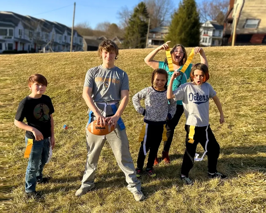 Children smiling and posing as one holds a football during a game of flag football outside of Ya